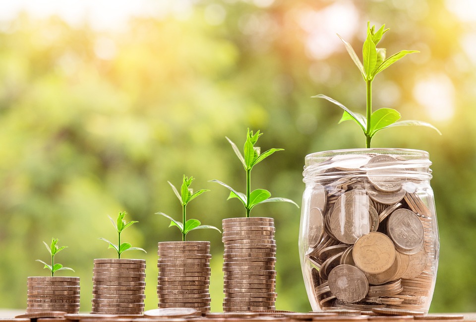 Stacks of coins with small plants shooting out the top of them. The stacks get taller from left to right, culminating in a full jar of cash.